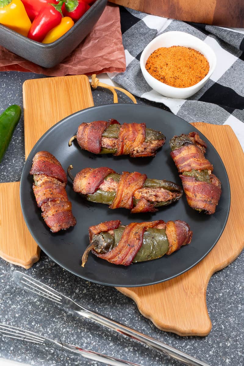Six bacon-wrapped brisket stuffed jalapeños are arranged on a black plate atop a Texas shaped wood cutting board. A bowl of seasoning, peppers, and utensils are nearby, creating a vibrant and appetizing food scene.