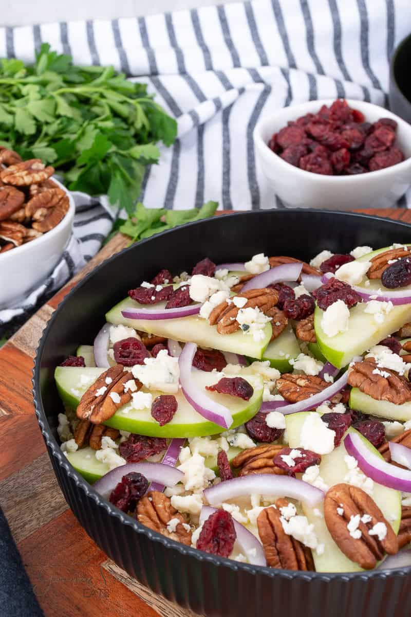 A black bowl filled with a salad of green apple slices, pecans, dried cranberries, red onion, and crumbled cheese. Fresh parsley, pecans, and dried cranberries are in small bowls nearby on a striped cloth.