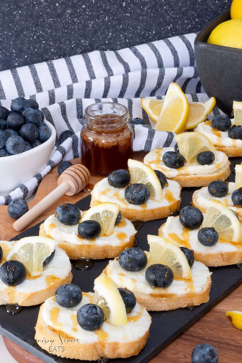 A slate board with bruschetta toast rounds topped with creamy lemon ricotta spread, fresh blueberries, and lemon wedges, drizzled with honey. A jar of honey, honey dipper, bowl of blueberries, and lemon slices are nearby. A striped towel is in the background.