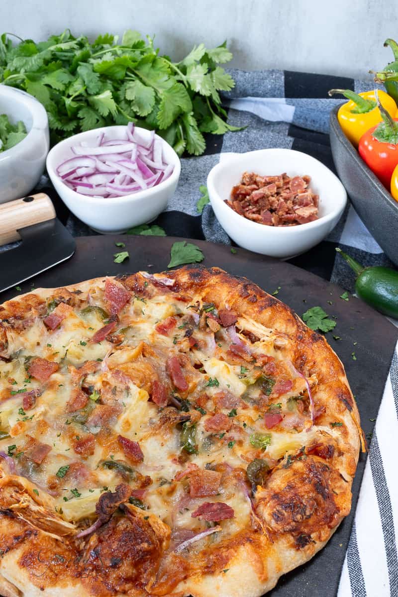 A baked pizza topped with cheese, bacon, and cilantro sits on a dark serving tray. Surrounding it are bowls of red onion and bacon, fresh cilantro, and colorful mini bell peppers on a tablecloth.
