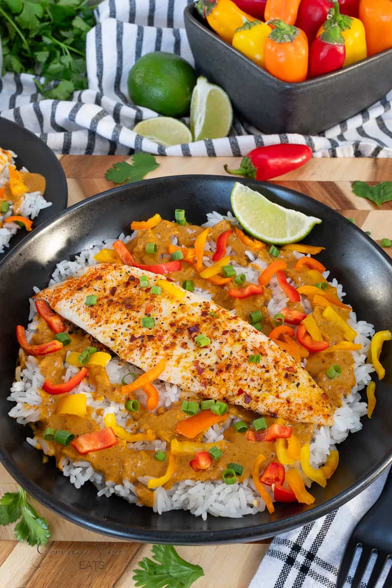 A black bowl with white rice topped with baked fish, creamy orange curry sauce, sliced bell peppers, and green onions, garnished with a lime wedge. Colorful mini peppers and limes are in the background.