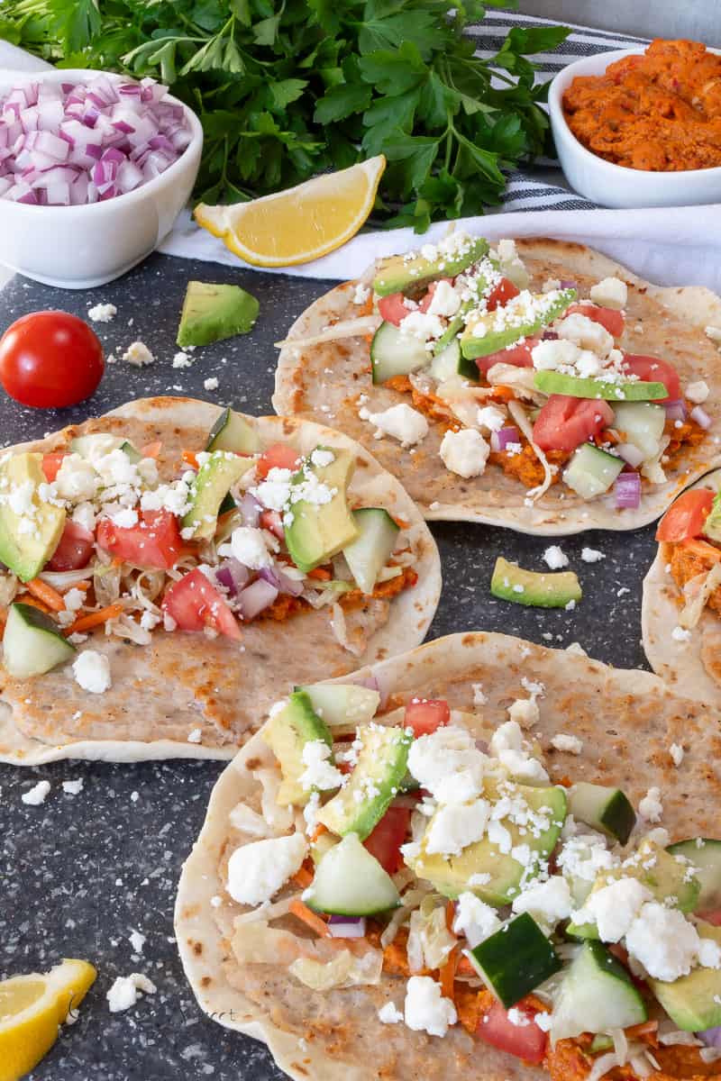 Four chicken smash tacos topped with shredded cabbage, diced cucumber, tomato, red onion, avocado, and crumbled cheese are displayed on a dark surface with lemon wedges, parsley, and bowls of chopped onions and sauce in the background.