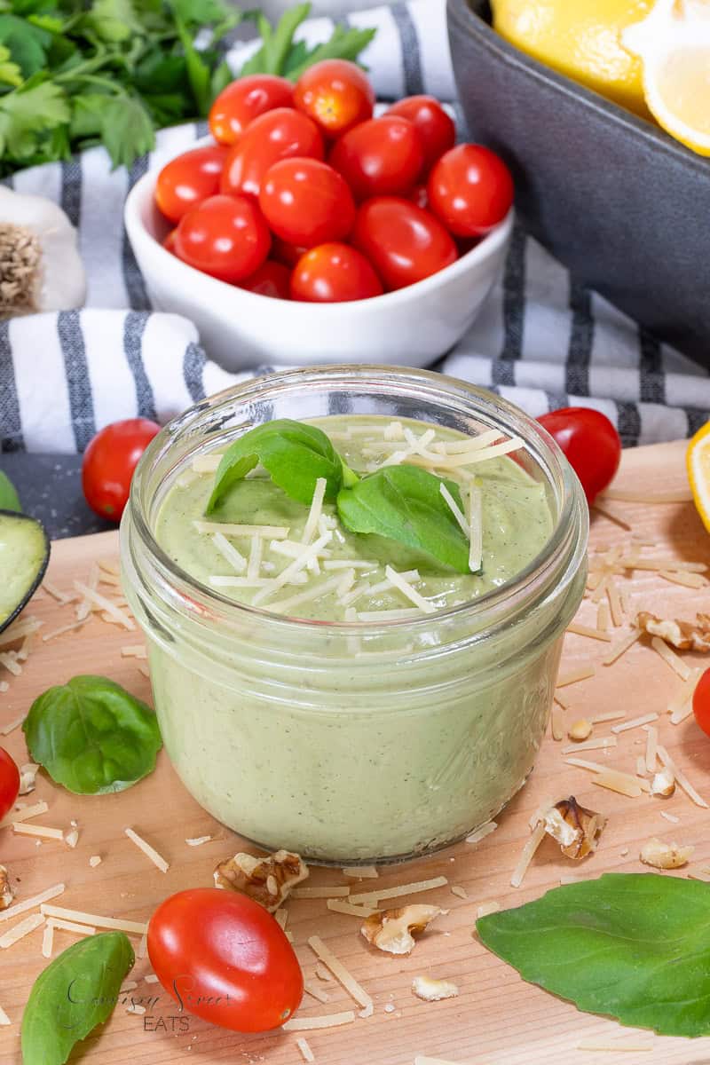 A glass jar filled with creamy green pesto topped with shredded cheese and basil leaves, surrounded by cherry tomatoes, basil, and walnuts on a wooden board. A bowl of cherry tomatoes and lemon slices are in the background.