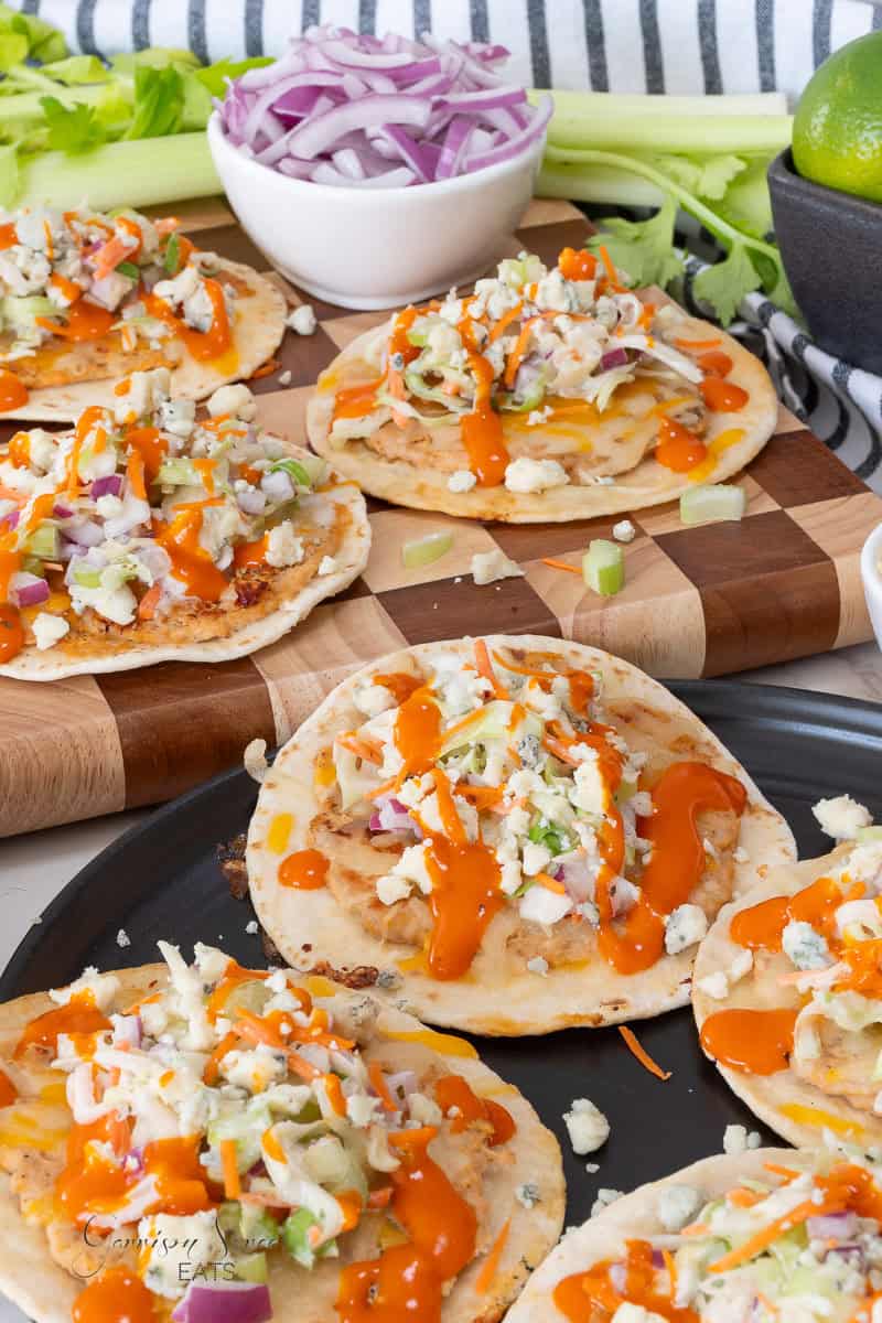 Small tortillas topped with shredded chicken, crumbled cheese, diced celery, chopped red onions, and drizzled with orange buffalo sauce, arranged on a cutting board and plate, with extra onions and limes in the background.