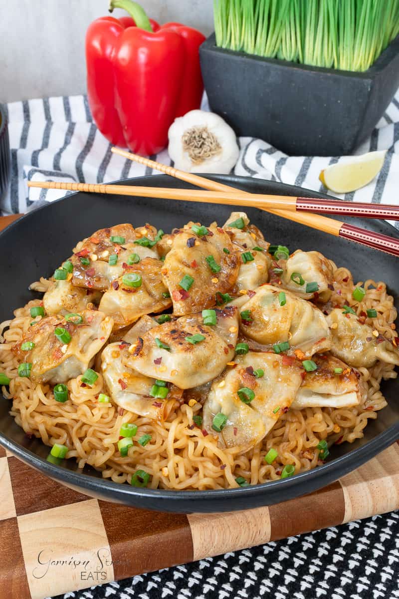 A black bowl filled with noodles topped with dumplings, chopped green onions, and chili flakes. In the background are a red bell pepper, garlic bulb, artificial grass, chopsticks, and a striped kitchen towel.