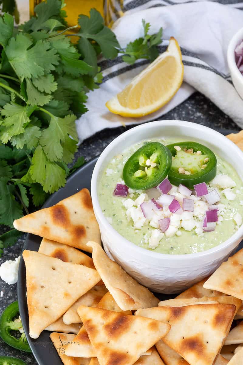 A bowl of creamy dip topped with jalapeño slices, chopped red onion, and crumbled cheese is surrounded by pita chips. Fresh cilantro and a lemon wedge are placed nearby on a dark surface.