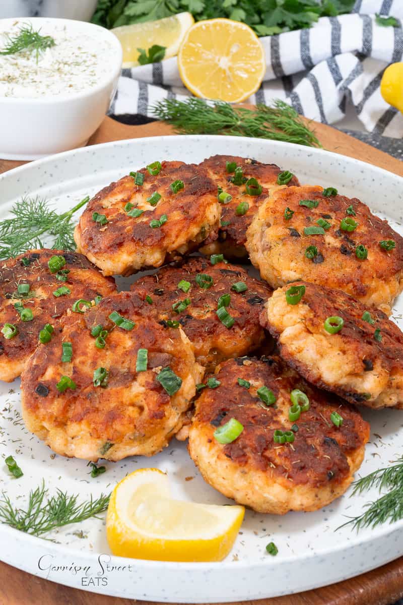 A plate of golden-brown salmon cakes garnished with chopped green onions and fresh dill, served with lemon wedges. A bowl of dipping sauce and a halved lemon are in the background.
