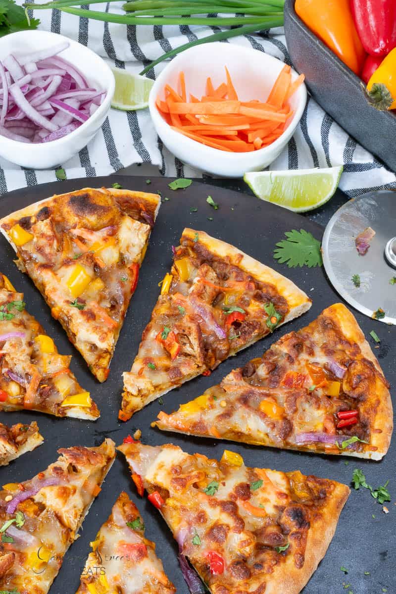 A sliced pizza topped with cheese, red onions, and yellow and red bell peppers on a black surface. Nearby are small bowls of sliced red onions and carrots, lime wedges, and a pizza cutter.