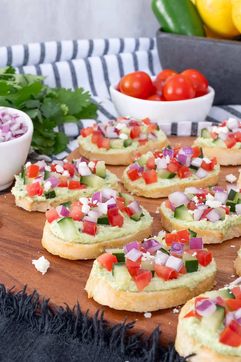 A wooden board with several pieces of toasted bread topped with creamy spread, diced tomatoes, cucumbers, red onions, and feta. Fresh vegetables and herbs are in the background.