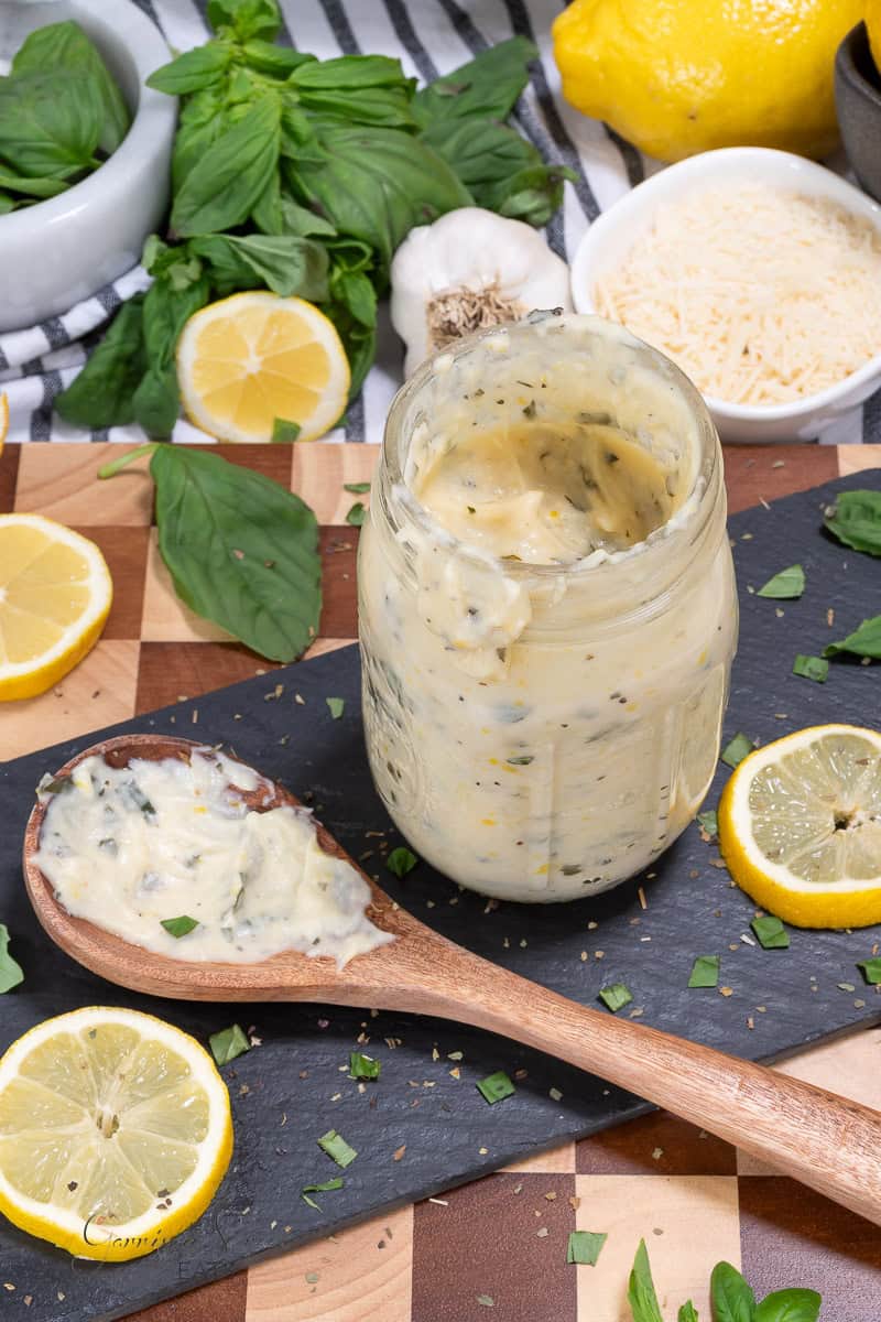 A jar of creamy lemon basil sauce sits on a slate board, surrounded by lemon slices, fresh basil leaves, grated cheese, and a wooden spoon topped with the sauce. Garlic and a lemon are in the background.