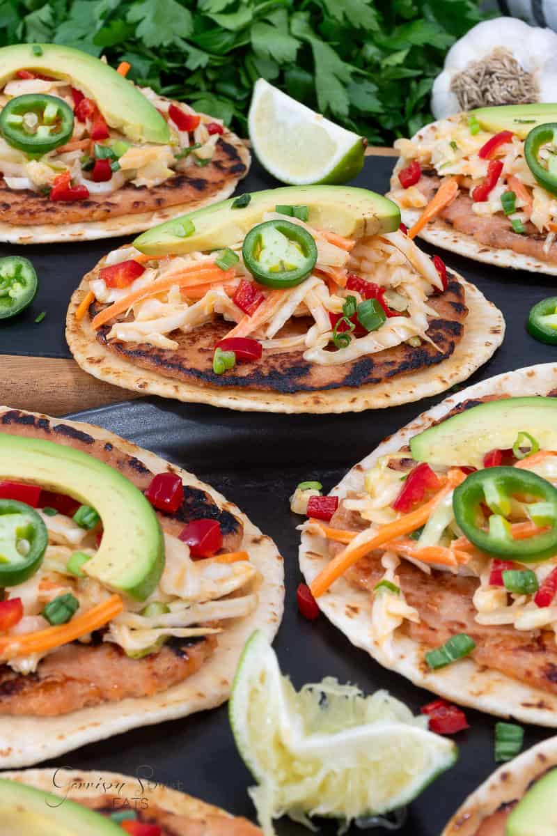 Grilled tortillas topped with smashed salmon, shredded cabbage, carrot, avocado slices, jalapeño, green onions, and red peppers, garnished with lime wedges. Fresh cilantro and a garlic bulb are in the background.