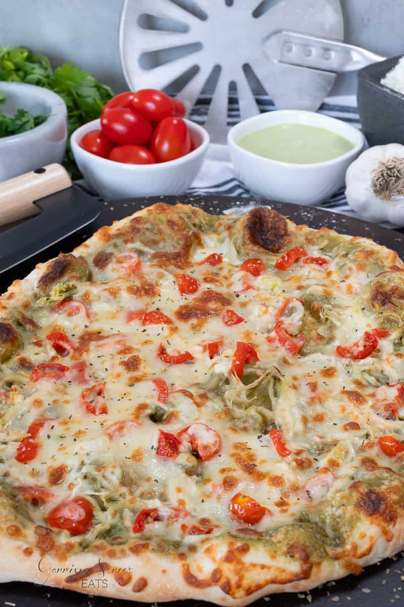 A homemade basil pesto chicken pizza topped with melted cheese, cherry tomato pieces, and green pesto sits on a black tray. Surrounding it are bowls of cherry tomatoes, green sauce, grated cheese, and fresh herbs.