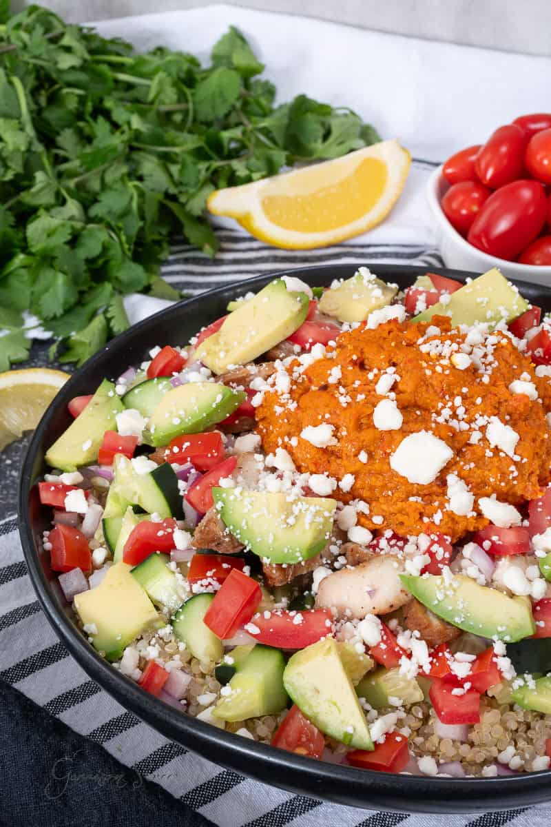 A bowl of quinoa salad with diced avocado, tomatoes, cucumber, onions, crumbled cheese, and a scoop of harissa hummus. Fresh cilantro, lemon wedges, and grape tomatoes are on the side.