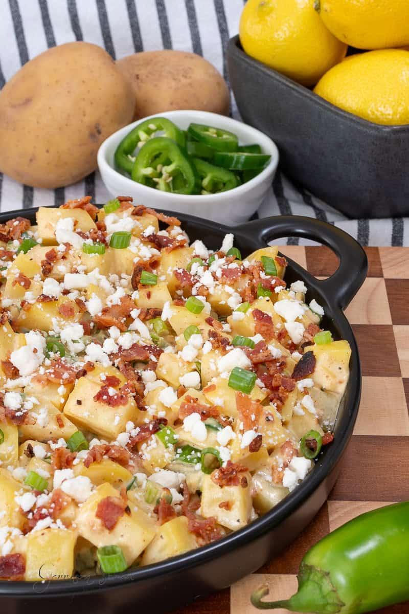 A black dish filled with potato salad topped with bacon, green onions, and crumbled cheese sits on a wooden board. Nearby are whole potatoes, sliced jalapeños, whole lemons, and a green chili pepper.