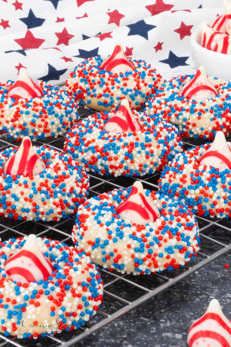4th of July blossom cookies topped with red and white striped candy kisses and coated in red, white, and blue sprinkles are cooling on a wire rack. A star-patterned cloth is in the background.