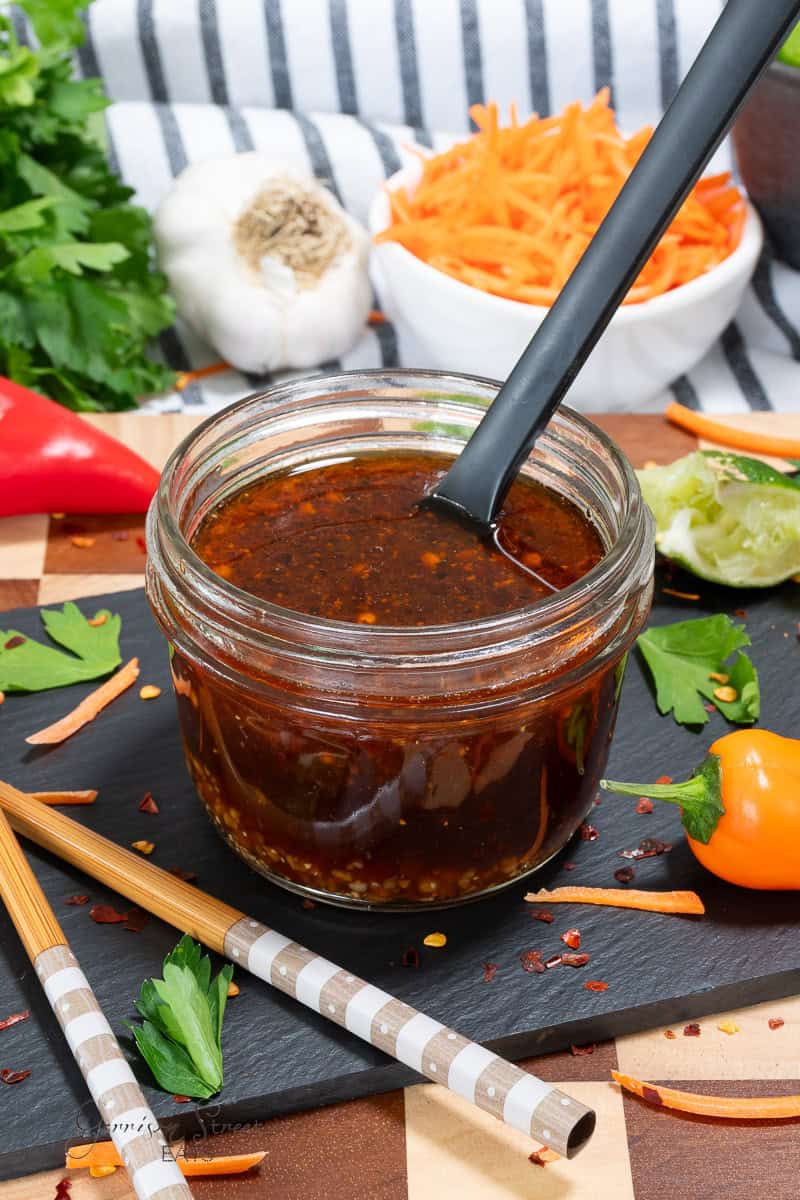 A glass jar filled with dark, spicy sauce and a spoon, surrounded by fresh parsley, sliced carrots, garlic, lime, a chili pepper, and chopsticks on a black slate board.