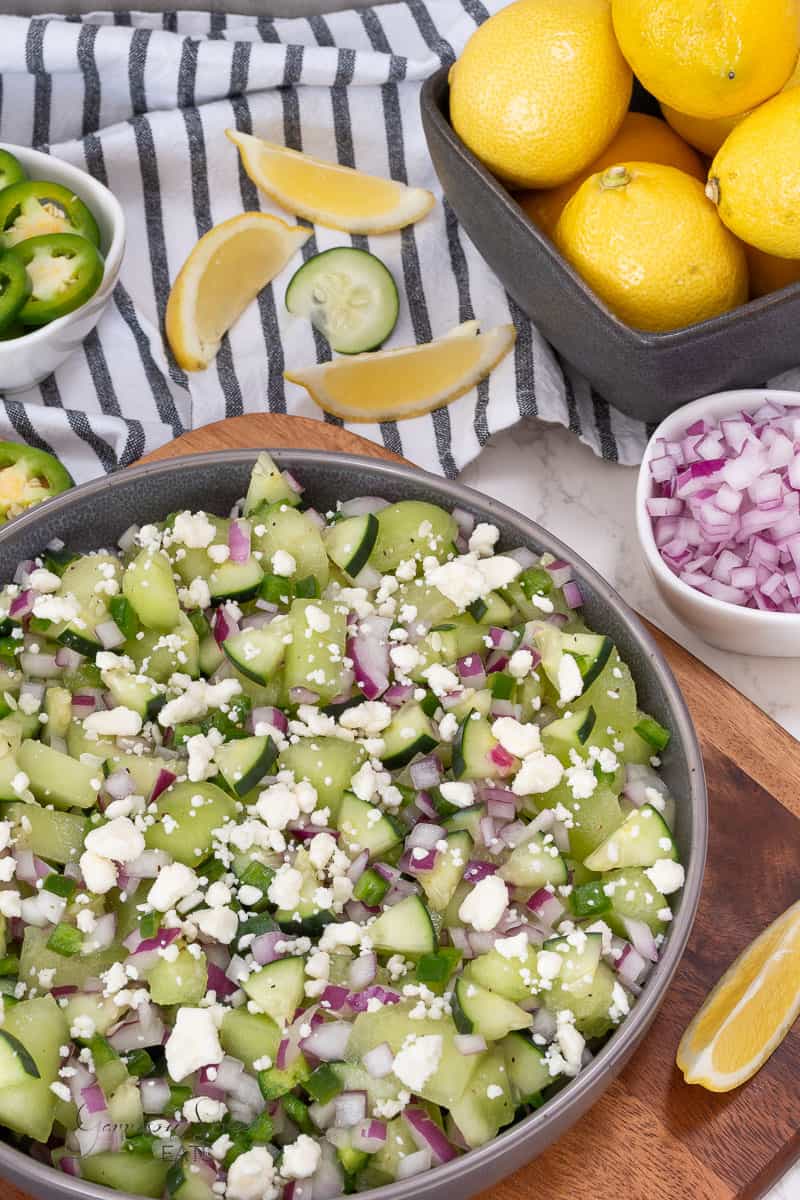 A bowl filled with honeydew salad topped with feta cheese and red onions sits on a wooden board. Nearby are lemon wedges, a bowl of whole lemons, sliced jalapeños, and a bowl of chopped red onions, with a striped towel in the background.
