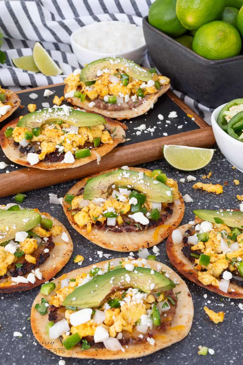 Crispy tortillas topped with scrambled eggs, sausage, avocado slices, crumbled cheese, and chopped onions and peppers, with limes and salsa on the side.
