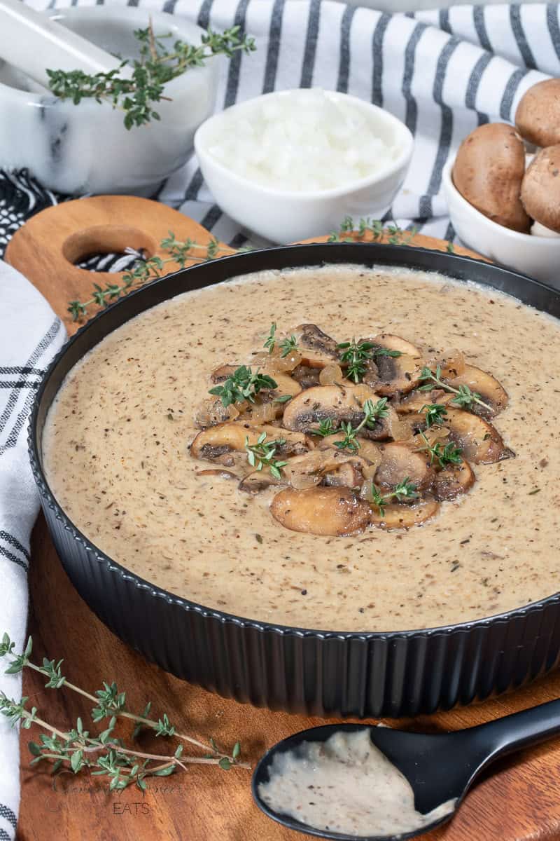 Creamy mushroom soup in a black bowl, garnished with sautéed mushrooms and fresh thyme. Nearby are a bowl of chopped onions, whole mushrooms, a marble mortar and pestle, and a black spoon with soup.