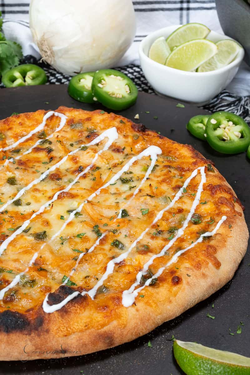 A baked pizza topped with melted cheese, herbs, and drizzled with white sauce, surrounded by sliced jalapeños, lime wedges, an onion, and fresh cilantro on a dark serving board.