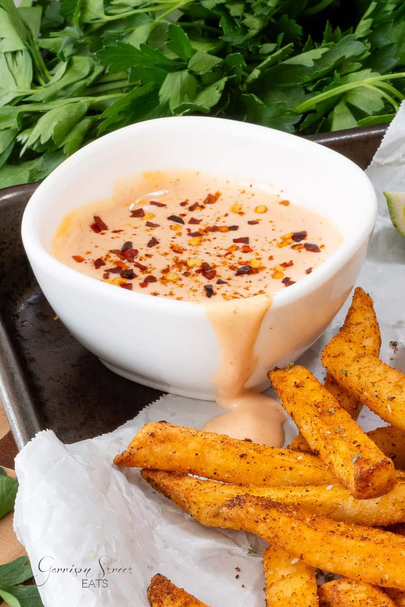 A white bowl of creamy dipping sauce topped with red pepper flakes sits next to seasoned fries on parchment paper, with fresh parsley in the background.