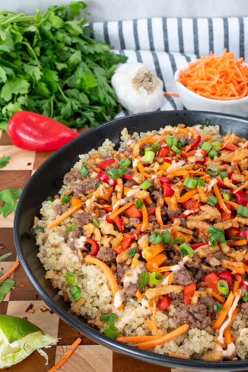 A bowl of quinoa topped with ground meat, shredded carrots, red pepper, crispy onions, and chopped green onions. Surrounding the bowl are fresh parsley, garlic, lime, bell pepper, and a bowl of shredded carrots.