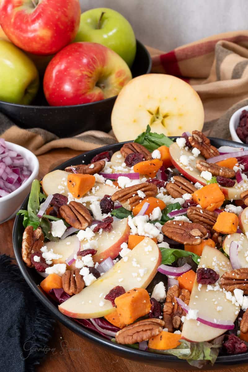 A salad with apple slices, pecans, sweet potatoes, feta cheese, red onions, and dried cranberries sits in a black bowl. Whole apples and small bowls of salad ingredients are in the background.