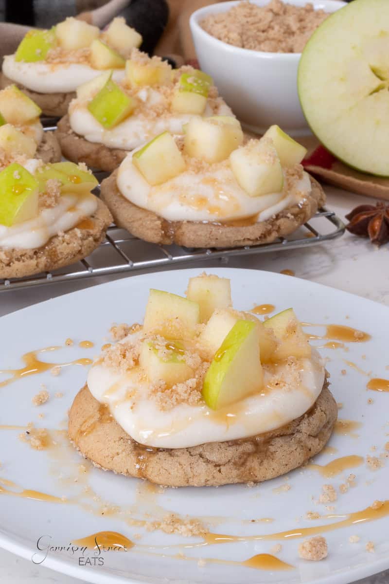 Close-up of cookies topped with white frosting, diced green apples, crumbly topping, and drizzled caramel sauce. More cookies and a bowl of crumble are in the background, with a sliced green apple on the side.