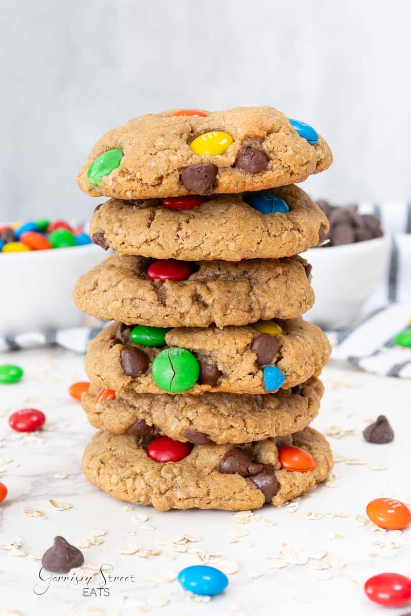 A stack of six oatmeal cookies with colorful candy-coated chocolates and chocolate chips, with more candies and a bowl in the background.