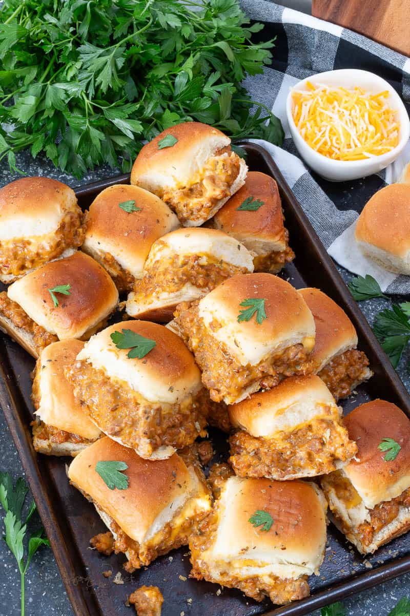 A tray of sloppy joe sliders on toasted rolls, topped with parsley leaves. A bowl of shredded cheese and fresh parsley are in the background.