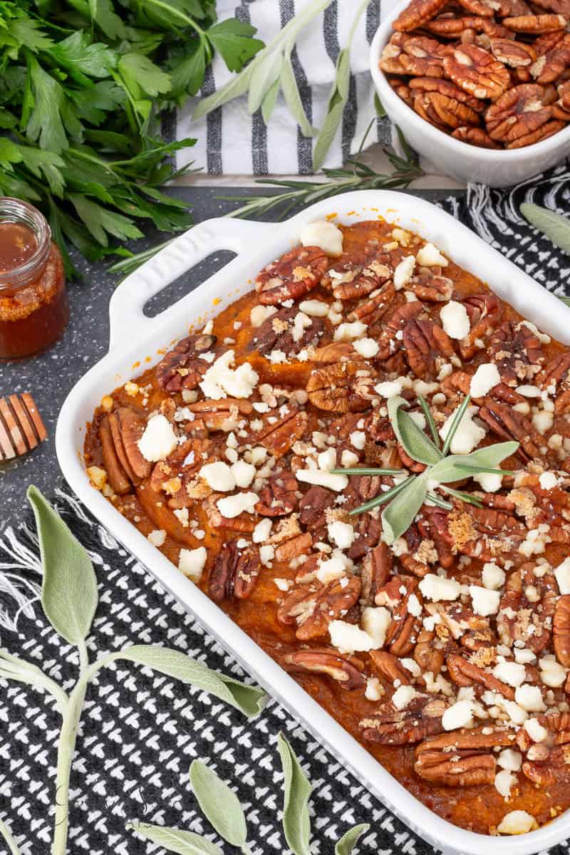 A baked sweet potato casserole topped with pecans and crumbled cheese sits in a white dish, surrounded by fresh herbs, a jar of honey, and a bowl of pecans on a dark patterned cloth.