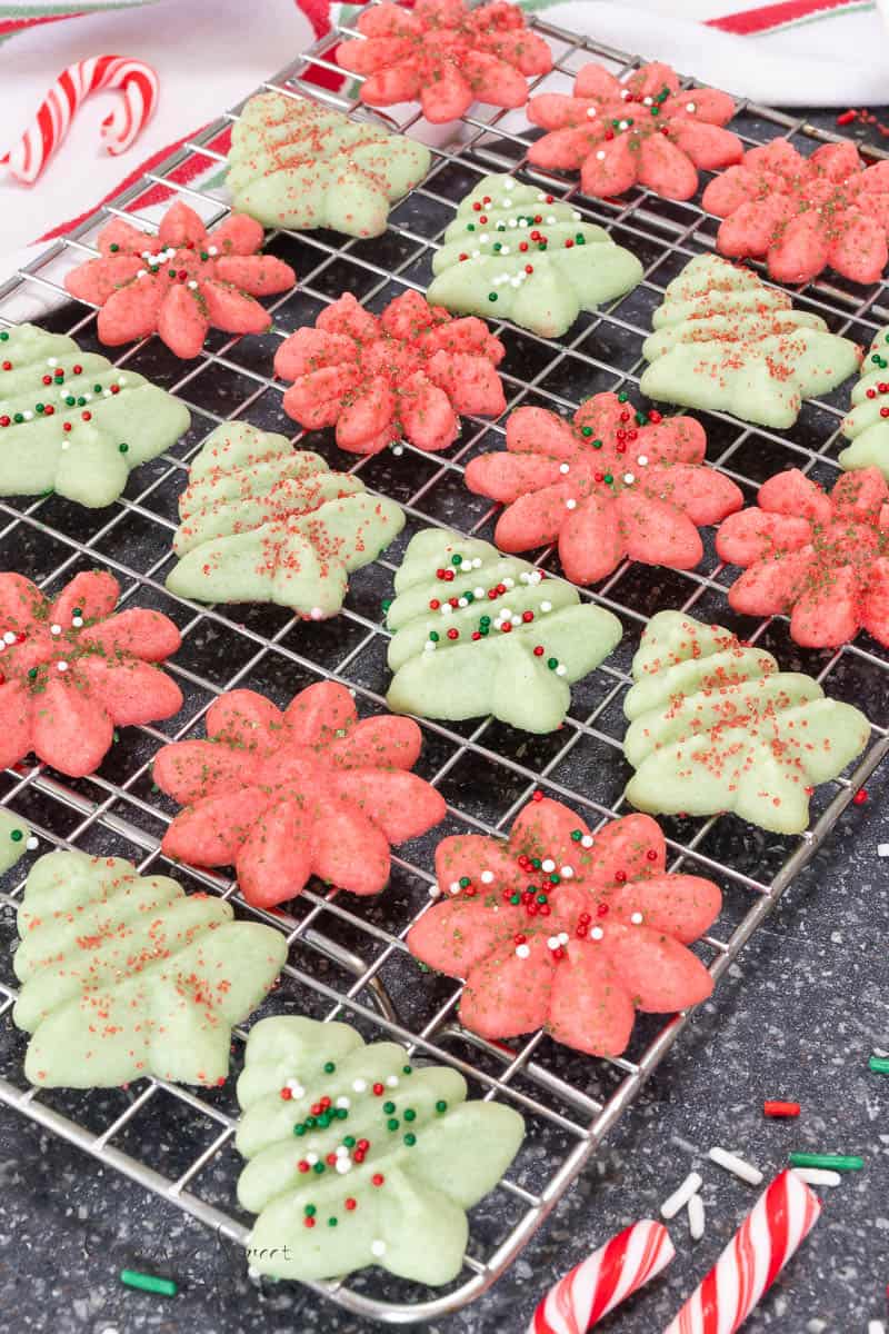 A wire cooling rack filled with red flower-shaped cookies and pale green Christmas tree cookies, both decorated with colorful sprinkles.