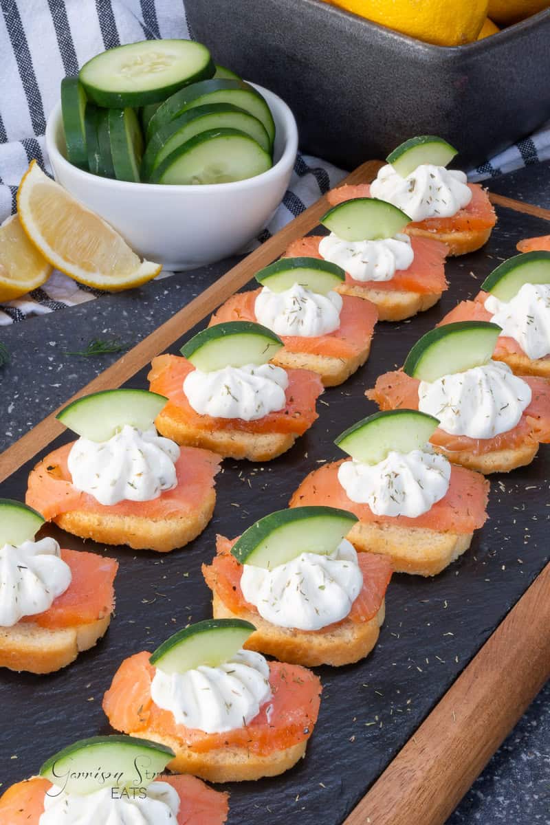 Smoked salmon canapés on toasted bread topped with cream cheese and cucumber slices, arranged on a slate serving board.