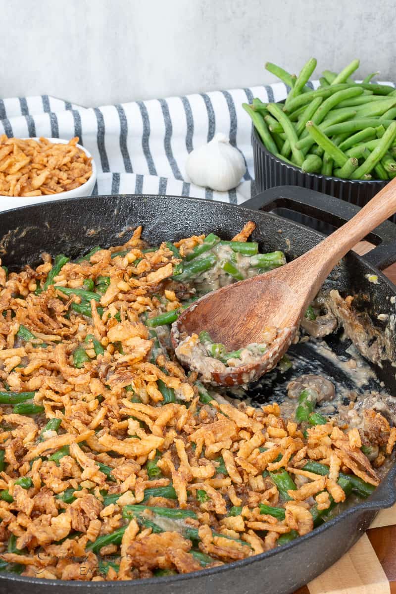Cast iron skillet of green bean casserole topped with crispy fried onions, served with a wooden spoon, fresh green beans, and a bowl of fried onions in the background.