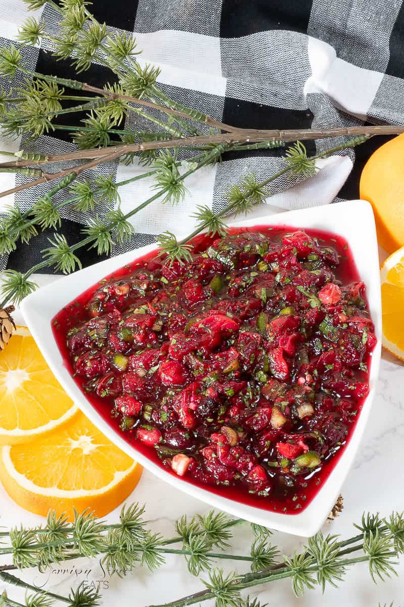 A triangular bowl of jalapeno cranberry sauce, garnished with orange slices and sprigs of greenery on a checkered towel.