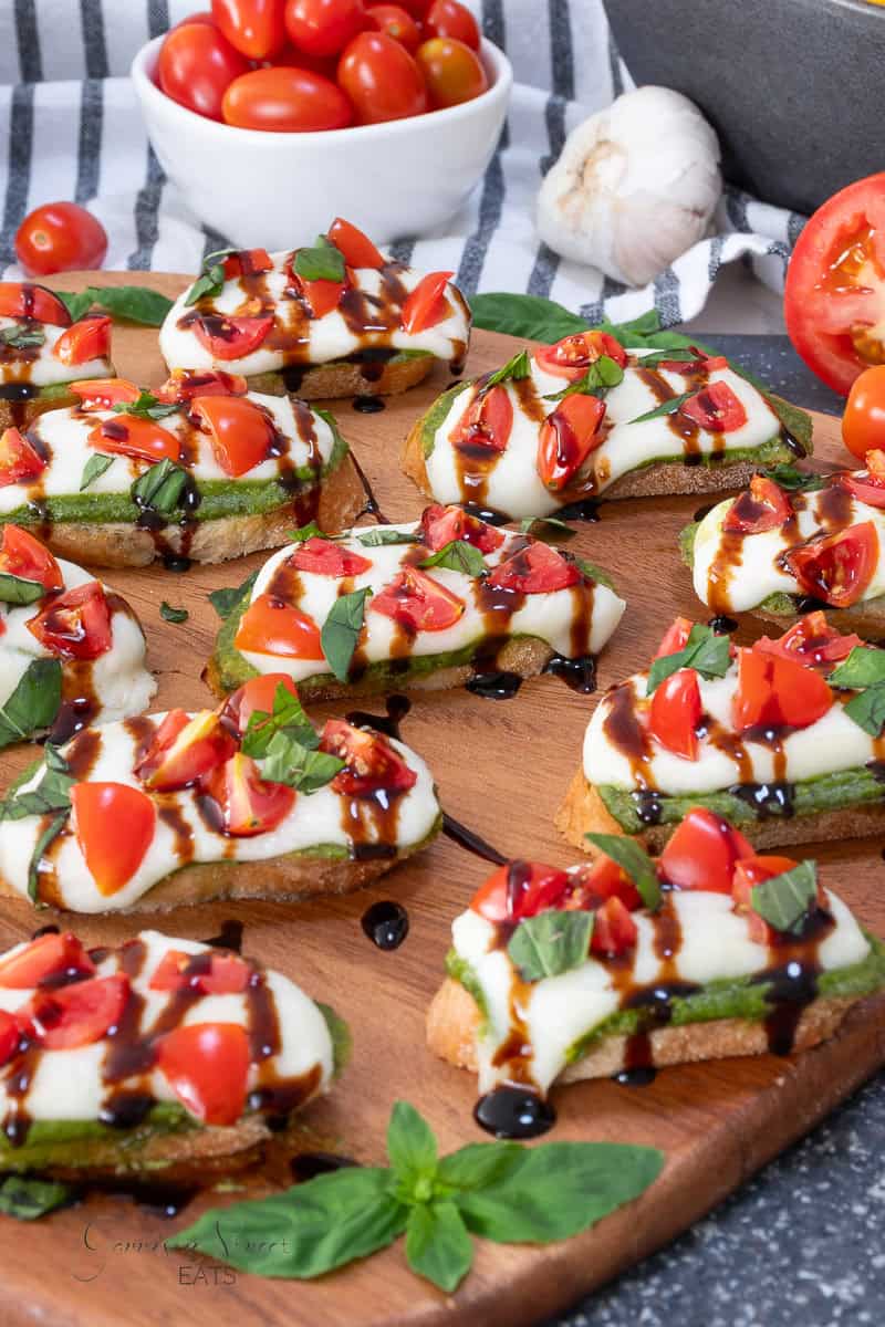 Several pieces of toasted bread topped with pesto, mozzarella, cherry tomatoes, and a balsamic glaze drizzle on a wooden board.