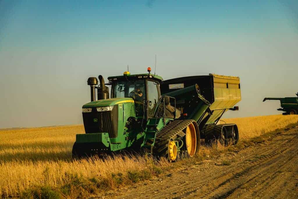 A green John Deere tractor with rubber tracks pulls a grain cart through a field of harvested Grasslands Canadian Khorasan under a clear sky, with a combine harvester working in the distance.