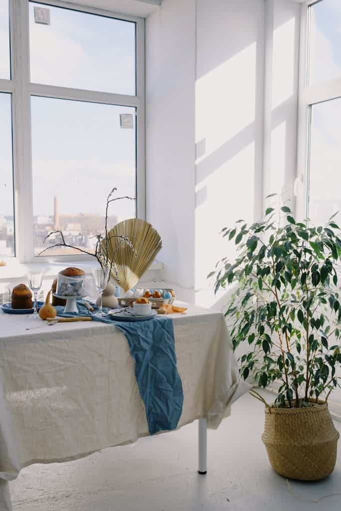 Bright table setting with a white tablecloth, ceramic ware, and indoor plants in sunlight.