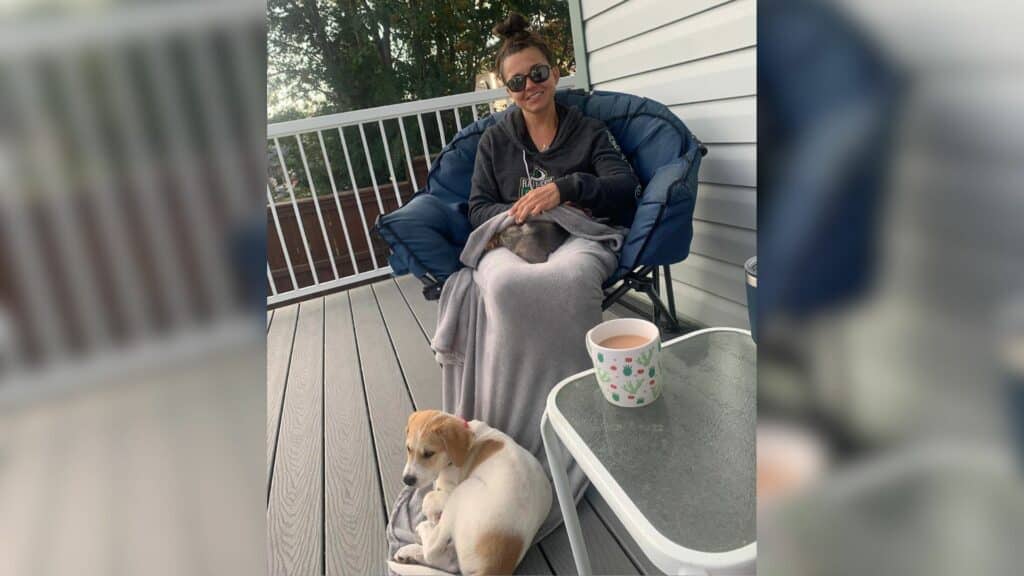 A woman with her hair pinned up and sunglasses on sitting in a camping chair with a blanket on her lap and a dog sitting in front of her.