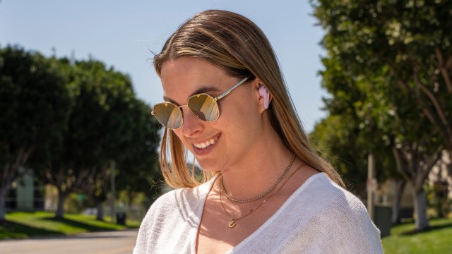 A young woman wearing sunglasses, looking at her phone in a leafy street with a pair of JBuds Pods in her ears