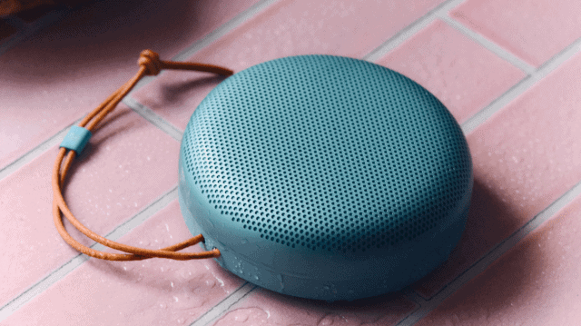 The Bang & Olufsen A1 3rd Gen in Eucalyptus Green on a pink bathroom tile, with water splashed over it