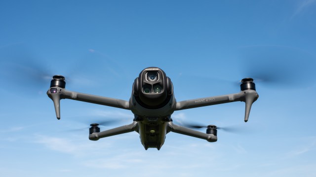 DJI Mavic 4 Pro in flight against a blue sky facing towards the camera with the drone camera and gimbal visible