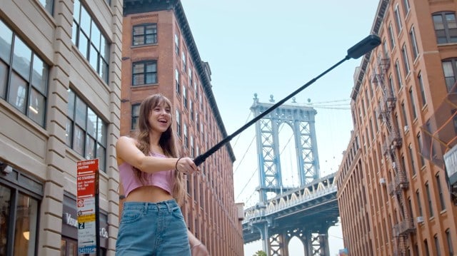 User holding a long selfie stick with Akaso 360 camera attached, Brooklyn bridge backdrop