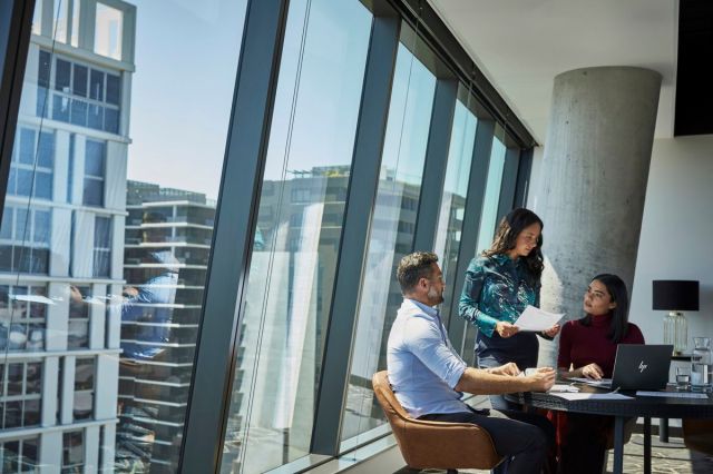 Three workers converse in an office. One uses the HP Elite Dragonfly G3.