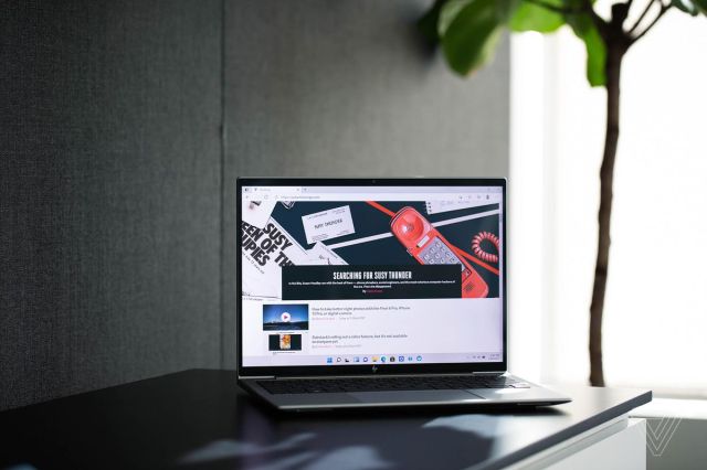 The HP Elite Dragonfly G3 on a table in front of a gray fabric wall. The screen displays The Verge homepage.