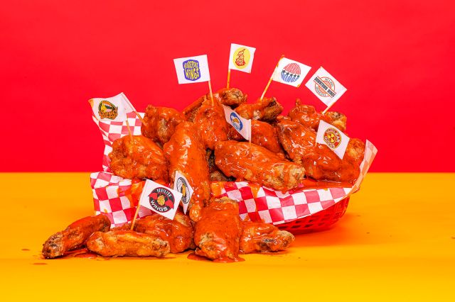 An image of a basket full of wings over a red and yellow background. Several wings have a toothpick planted in them connected to a flag with a different made up chicken wing restaurant logo. 