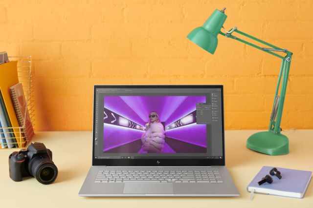 The HP Envy 17 on a desk, open. The screen displays a photo editing software. On its left is a camera and a bin of notebooks, on its right are a notebook, a pair of earbuds, and a lamp.