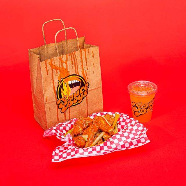 A bag with a sticker for a fake brand called Sauce Boys next to a basket of chicken wings and a clear plastic cup with another sticker for Sauce Boys on a red backdrop.