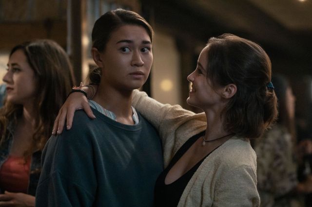 A woman has her arm around the neck of another woman and looks at her fondly while the other woman looks nervously away.