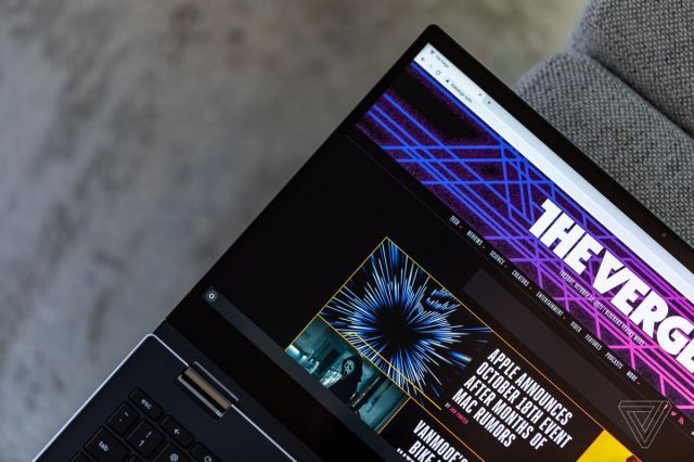 The Asus Chromebook Flip CX5 laid out flat on a gray fabric bench. The screen displays The Verge homepage.